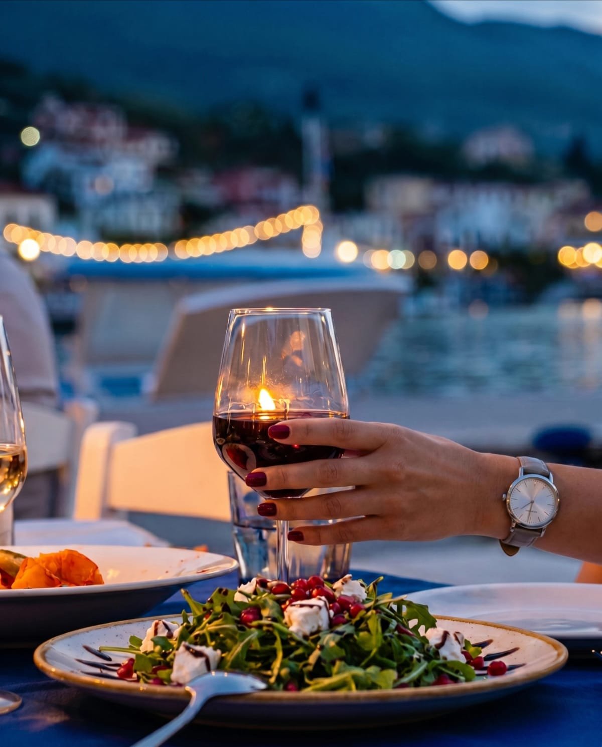 A glass of red wine held above a harbor-side table at dusk.